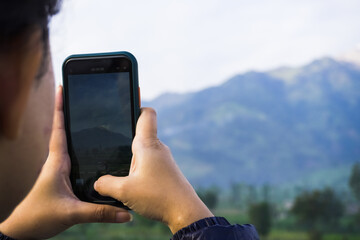 A woman wearing goose down jacket is taking a photo of a mountain and plantation landscape in the morning using her smartphone. Merapi Merbabu Mountainside in Selo Boyolali, Central Java, Indonesia.