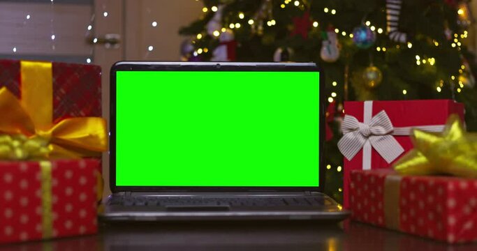 Laptop with green screen surrounded by gifts on background of Christmas tree