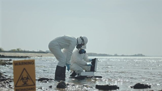 Two ecologists wearing protective coveralls and respiratory masks collecting samples of lake water in polluted area with warning biohazard sign