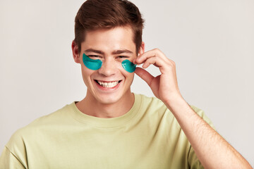 Young handsome smiling guy with brown hair applying under eye hydrogel patches. Caucasian millennial man practices skin care routine to keep healthy and youthful looking. Studio portrait.
