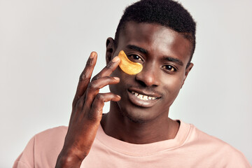 Young handsome black guy applying cosmetic under eye hydrogel patch on his face. African American millennial man practices skin care routine to keep healthy and youthful looking. Studio portrait.