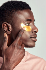 Close up portrait of young handsome black guy applying cosmetic under eye hydrogel patch on his face. African American millennial man practices skin care routine to keep healthy and youthful looking.