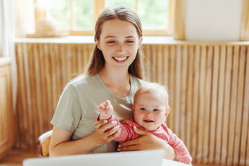 Smiling young mother holding beautiful cute baby, using laptop, video conference, working online