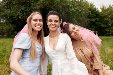 Happy adorable bride in a white dress with white veil and two bridesmaids in blue and beige dress with pink veils sitting on the grass in a summer park hugging each other. Cheerful bachelorette party.