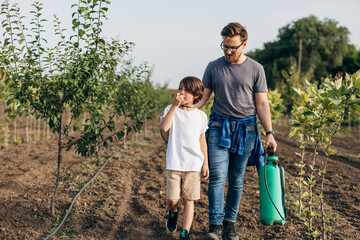 Father and son walking trough the orchard with a sprayer.