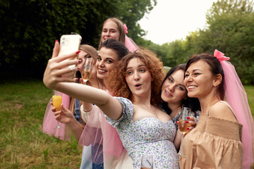 Happy charming bride in a white dress and bridesmaids with pink veils hug and take a selfie holding champagne glasses in their hands. Cheerful bachelorette party in the summer park.
