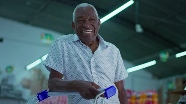One happy Brazilian senior shopper smiling at camera with shopping cart inside supermarket shed. Portrait of an African American older consumer at grocery store