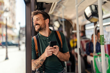 An urban commuter is standing in a public bus and looking through the window while typing on the phone.