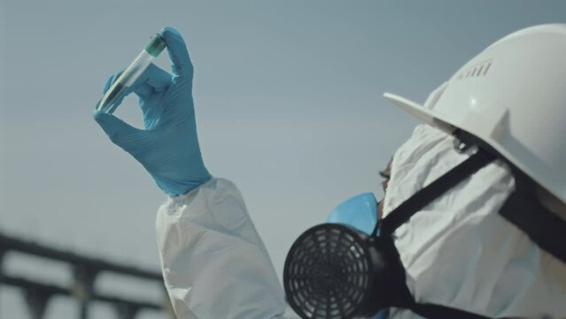 Low angle shot of ecologist in protective coverall, gloves and respiratory mask holding test tube and examining sample