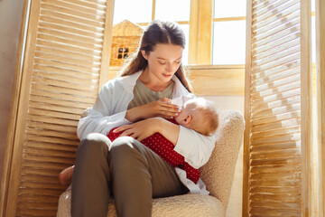 Young mother breastfeeding her cute little baby in bedroom at home. Woman holding child in her arms