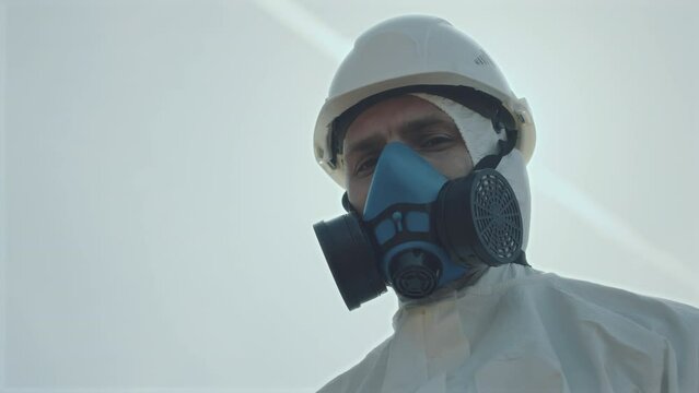 Low angle portrait shot of male ecologist in protective coverall and respiratory mask standing outdoors and posing for camera