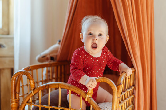 Adorable, happy child standing in wooden playpen looking at camera in bedroom at home