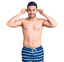 Fototapeta premium Young handsome man wearing swimwear smiling pulling ears with fingers, funny gesture. audition problem