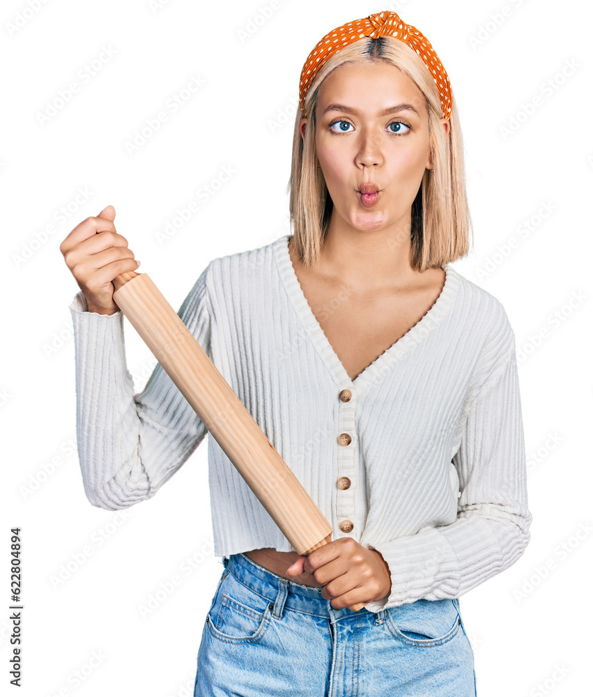 Poster beautiful young blonde woman holding kneading roll making fish face with mouth and squinting eyes, c