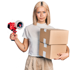 Beautiful young blonde woman holding packing tape and cardboard boxes skeptic and nervous, frowning upset because of problem. negative person.