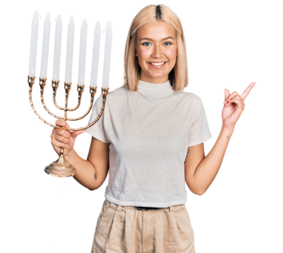 Beautiful young blonde woman holding menorah hanukkah jewish candle smiling happy pointing with hand and finger to the side