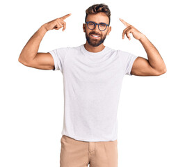 Young hispanic man wearing casual clothes and glasses smiling pointing to head with both hands...