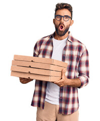 Young hispanic man holding delivery pizza box scared and amazed with open mouth for surprise,...