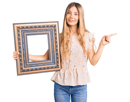 Beautiful caucasian woman with blonde hair holding empty frame smiling happy pointing with hand and finger to the side