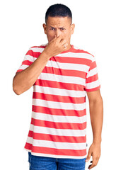 Young handsome latin man wearing casual clothes smelling something stinky and disgusting, intolerable smell, holding breath with fingers on nose. bad smell