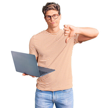 Young handsome man wearing glasses holding laptop with angry face, negative sign showing dislike with thumbs down, rejection concept