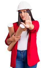 Young beautiful brunette woman wearing hardhat holding architect blueprint with open hand doing stop sign with serious and confident expression, defense gesture