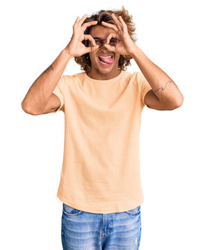 Young Hispanic Man Wearing Casual Clothes And Glasses Doing Ok Gesture Like Binoculars Sticking Tongue Out, Eyes Looking Through Fingers. Crazy Expression.