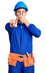 Young handsome man wearing worker uniform and hardhat laughing at you, pointing finger to the camera with hand over mouth, shame expression