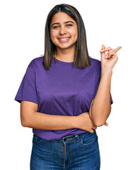 Young hispanic girl wearing casual purple t shirt with a big smile on face, pointing with hand and finger to the side looking at the camera.