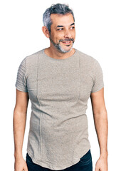 Middle age hispanic with grey hair wearing casual grey t shirt looking away to side with smile on face, natural expression. laughing confident.