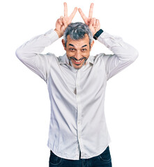 Middle age hispanic with grey hair wearing casual white shirt posing funny and crazy with fingers on head as bunny ears, smiling cheerful