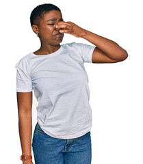 Young african american woman wearing casual white t shirt smelling something stinky and disgusting,...