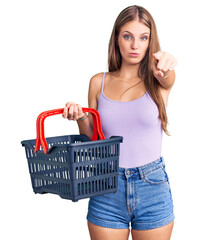 Young beautiful blonde woman holding supermarket shopping basket pointing with finger to the camera and to you, confident gesture looking serious