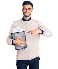 Young handsome hispanic man holding paper bin full of crumpled papers smiling happy pointing with hand and finger