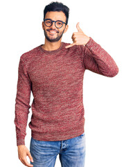 Young handsome hispanic man wearing winter sweater and glasses smiling doing phone gesture with...