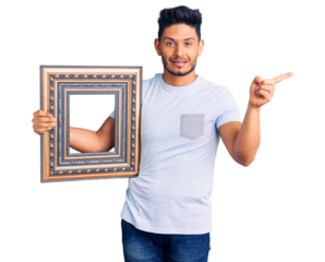 Handsome latin american young man holding empty frame smiling happy pointing with hand and finger to the side