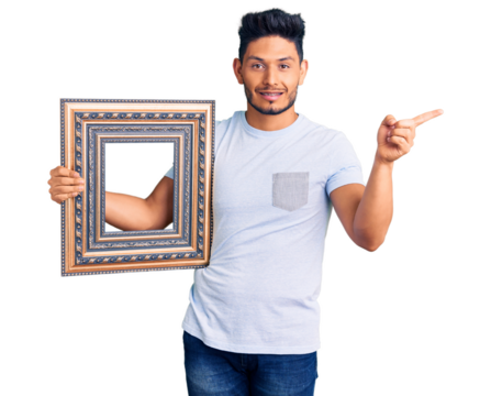 Handsome latin american young man holding empty frame smiling happy pointing with hand and finger to the side