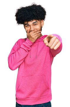 Young african american man with afro hair wearing casual pink sweatshirt laughing at you, pointing finger to the camera with hand over mouth, shame expression