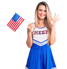 Young beautiful blonde woman wearing cheerleader uniform holding united states flag doing ok sign with fingers, smiling friendly gesturing excellent symbol