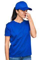 Young hispanic girl wearing delivery courier uniform smelling something stinky and disgusting,...