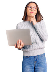 Young beautiful chinese girl wearing glasses holding laptop serious face thinking about question...