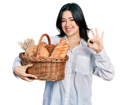 Beautiful hispanic woman holding wicker basket with bread doing ok sign with fingers, smiling friendly gesturing excellent symbol