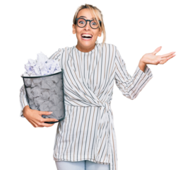 Beautiful blonde business woman holding paper bin full of crumpled papers celebrating victory with happy smile and winner expression with raised hands