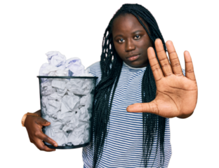 Young black woman with braids holding paper bin full of crumpled papers with open hand doing stop sign with serious and confident expression, defense gesture