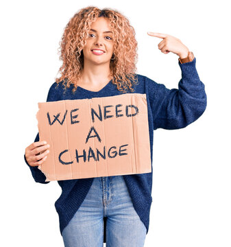 Young blonde woman with curly hair holding we need a change banner pointing finger to one self smiling happy and proud