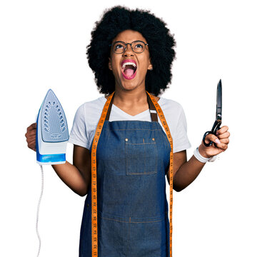 Young african american woman dressmaker designer wearing atelier apron holding iron and scissors angry and mad screaming frustrated and furious, shouting with anger looking up.