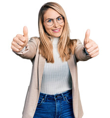 Young blonde woman wearing business shirt and glasses approving doing positive gesture with hand, thumbs up smiling and happy for success. winner gesture.