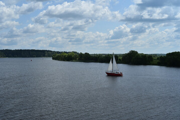 A sailboat floats down the river with people on board. In the background, a beautiful wooded shore, a turn of the river, a cloudy sky.