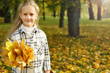 Little blonde girl posing in a white plaid shirt in an autumn park with a bouquet of yellow leaves