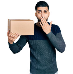 Young arab man with beard holding delivery package covering mouth with hand, shocked and afraid for...